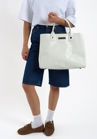 Witte tas met krokodillenprint en een gestructureerd ontwerp, met twee stevige handvatten en een textuurafwerking, vastgehouden door iemand die denim shorts draagt.