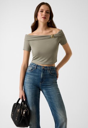 Femme portant un haut olive vert décolleté, un jean bleu taille haute, des boucles d'oreilles dorées, tenant un sac à main noir sur un fond blanc.