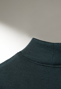 Mörkgrön ribbad polokrage med vertikal struktur. Sömdetaljer synliga; tyget verkar mjukt och stretchat.