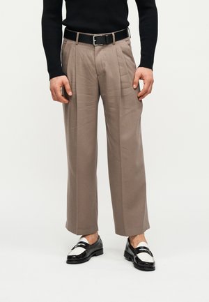 Homme portant un pantalon plissé beige avec une ceinture noire, des mocassins noirs et blancs, et un haut à manches longues noir, debout devant un fond blanc.