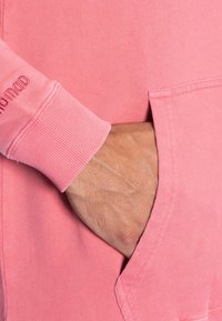 Roze sweatshirt met ribgebreide manchetten en een voorkantzak, in close-up weergegeven met een hand die in de zak rust. Zachte stofstructuur zichtbaar.
