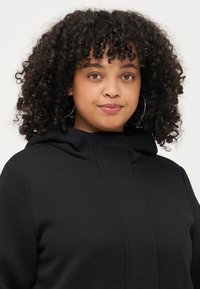 Sweat-shirt à capuche noire avec une texture douce, présentant une fermeture éclair avant et un design minimaliste. Tissu épais avec une coupe confortable.