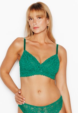 Femme blonde portant un soutien-gorge en dentelle verte et une lingerie assortie, posant avec une main sur la hanche sur fond blanc.