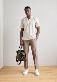 Camisa blanca de manga corta con textura y dos bolsillos en el pecho, combinada con pantalones marrones claros y una bolsa verde oscura con detalles marrones. Zapatillas blancas.
