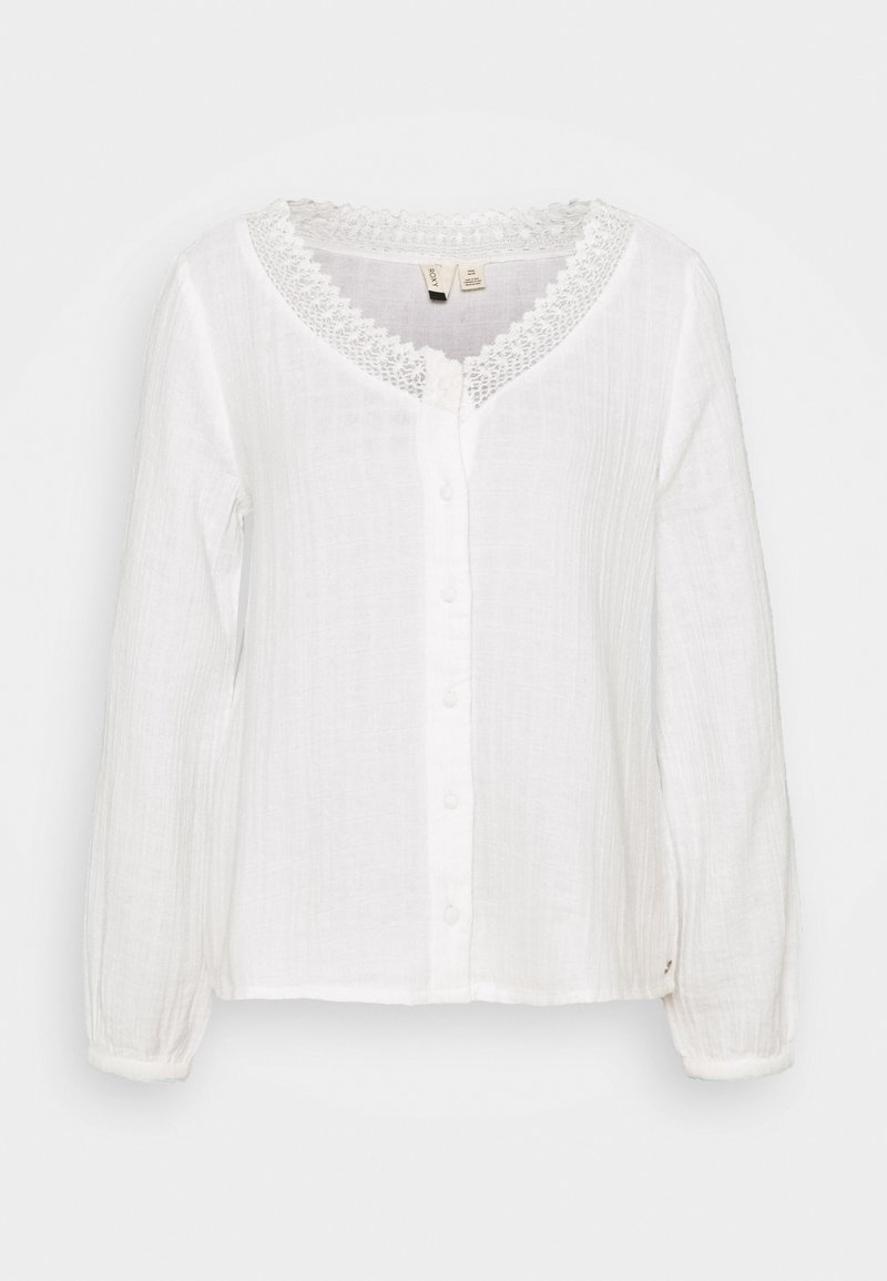 Blouse blanche à manches longues en tissu texturé avec un col rond bordé de dentelle, fermeture boutonnée à l'avant et poignets élastiqués.