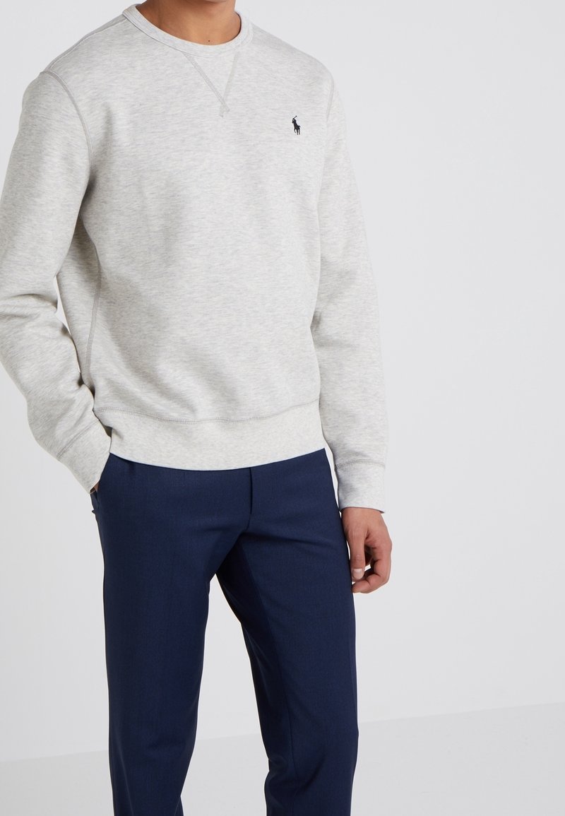 Felpa grigia chiaro con scollo rotondo e polsini a costine, dotata di un piccolo logo nero. Abbinata a pantaloni blu navy sartoriali.