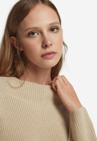 Beige gestrickter Pullover mit gerippter Textur und weitem Halsausschnitt, getragen von einer Person mit langen Haaren, die die Stoffdetails des Kleidungsstücks zeigt.