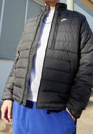 Person trägt schwarze Nike Steppjacke über weißem Hemd und blauen Hosen, steht draußen in der Nähe eines Gebäudes mit geschlossenen Fensterläden.