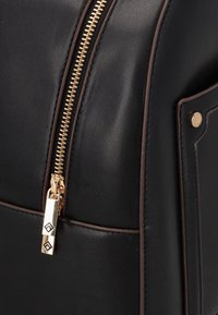 Detailní záběr na černou koženou tašku se zlatým zipem a tmavě hnědými lemy, který zdůrazňuje táhlo zipu a detaily kovových nýtků.