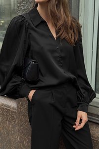 Black satin button-up shirt with puffed sleeves, paired with tailored black trousers. Small black bag with a chain strap. Smooth texture.