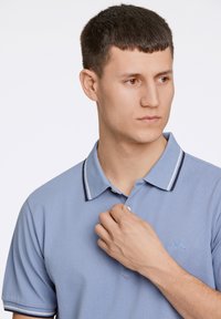 Lyseblå polo skjorte med en tekstureret stof, en kontrasterende krave med navy- og hvide striber samt broderet logo på brystet.