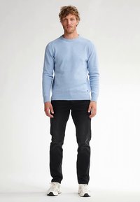Hellblauer Strickpullover mit gerippten Bündchen und Saum, kombiniert mit schwarzen Jeans und weißen Sneakers. Das Modell hat lockiges Haar und steht aufrecht.