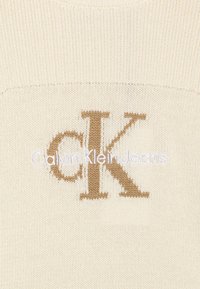 Maglione a maglia color crema con finitura testurizzata, caratterizzato da un grande logo 'CK' ricamato in marrone e 'Calvin Klein Jeans' in bianco.