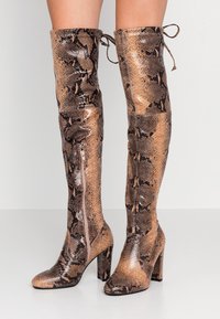 Bottes hautes jusqu'à la cuisse à talons hauts avec un motif de peau de serpent marron et noir et des fermetures éclair sur les côtés, portées sur des jambes nues sur un fond neutre.