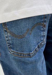 Poche arrière d'un jean en denim bleu avec des coutures orange, partiellement recouverte par un vêtement blanc.