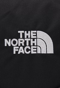 Čierna tkanina so vyšívaným bielym logom s textom "THE NORTH FACE" a zakriveným polkruhovým dizajnom. Textúrovaný povrch, minimalistický dizajn.