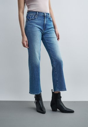 Žena nosí modré rifle s vysokým pásom a rovnými nohavicami a čierne členkové topánky, stojí na jednoduchom svetlosivom pozadí.