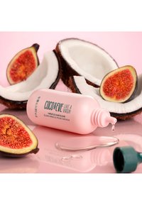 Roze fles haarelixer met groene dop, gepositioneerd nabij gespleten kokosnoten en vijgen op een roze oppervlak. Duidelijke vloeistofdruppel in de buurt.