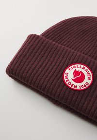 Mørkerød ribbet strikknisse med et rundt hvidt og rødt Fjällräven Sverige 1960 logo-patch på kanten.
