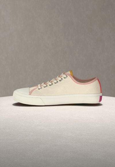 Χαμηλό μπεζ αθλητικό παπούτσι με λευκή ελαστική σόλα, μεταλλικά ροδέλες, ροζ λεπτομέρειες και κόκκινη ετικέτα στη φτέρνα, τοποθετημένο σε φωτεινή επιφάνεια με μπεζ φόντο.