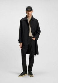 Zwarte waterdichte trenchcoat met dubbele knopen, gedragen over een beige shirt, gecombineerd met donkere spijkerbroek en zwarte sneakers met een crèmekleurige zool.