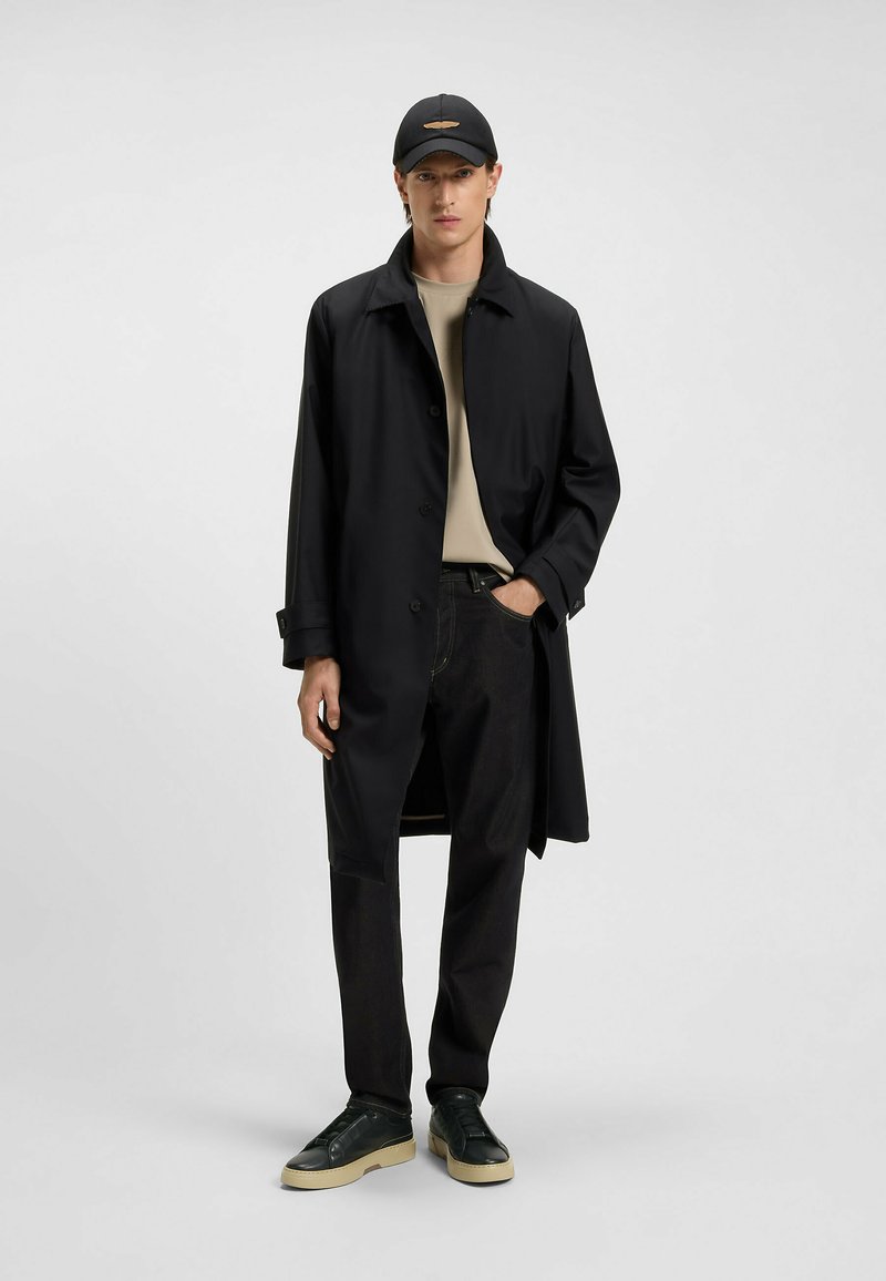 Zwarte waterdichte trenchcoat met dubbele knopen, gedragen over een beige shirt, gecombineerd met donkere spijkerbroek en zwarte sneakers met een crèmekleurige zool.