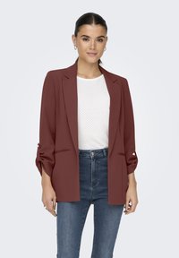 Bordeaux blazer met op maat gemaakte revers, twee voorkruiszakken en opgerolde mouwen, gecombineerd met een witte structuurblouse en blauwe jeans.