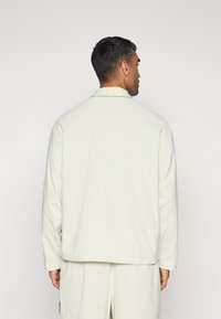 Light cream long-sleeve shirt with a collared neck and textured fabric, featuring a straight hem and subtle stitching details.