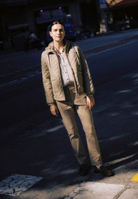 Mlada ženska stoji na sončni prehodu za pešce na sončni ulici, oblečena v bež prešito jakno, črtasto majico, svetle hlače in temne čevlje.