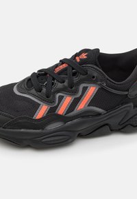 Mustat urheilukengät, joissa on verkkomateriaali päällinen, musta ja oranssi korostus sekä kuvioitu kumipohja, jossa on erottuva aaltomainen muotoilu.