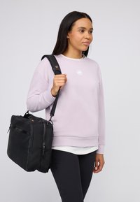 Kvinna iklädd en ljuslila sweatshirt och svarta byxor, bärande en svart axelväska, stående mot en enkel ljus bakgrund.