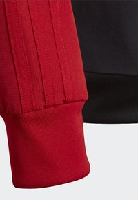 Red sweatshirt with a ribbed cuff and vertical seam details, paired with a black fabric section. Textured cotton blend material.