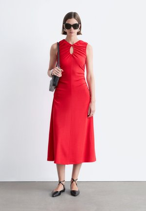 Kobieta w czerwonej, bezrękawnikowej sukience midi z dekoltem w kształcie klucza, czarnych spiczastych balerinach, czarnej torebce na ramię i dużych czarnych okularach przeciwsłonecznych.