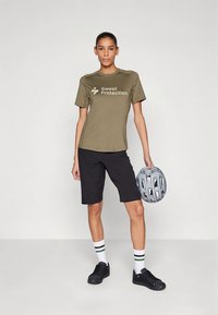 Alyvuogių žalios spalvos drėgmę sugeriantis marškinėlis su "Sweet Protection" logotipu, derinamas su juodais šortais, baltais kojiniais su žaliais dryžiais ir juodais sportbačiais, laikant pilką šalmą.