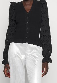 Schwarze Strickweste mit floral bestickten Ärmeln, V-Ausschnitt, Knopfleiste, gerafftem Saum, kombiniert mit glänzenden silbernen Hosen.