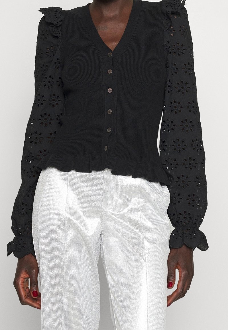 Schwarze Strickweste mit floral bestickten Ärmeln, V-Ausschnitt, Knopfleiste, gerafftem Saum, kombiniert mit glänzenden silbernen Hosen.