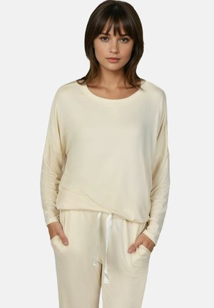 Jeune femme debout face à l’avant, portant un haut ample crème à manches longues et un pantalon avec cordon assorti, les mains dans les poches.