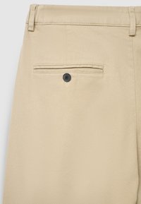 Dettaglio posteriore dei pantaloni beige su misura con una tasca a filetto singola chiusa da un bottone nero e passanti per cintura sulla vita.