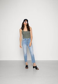 Top de malha verde, jeans de cintura alta azul claro com ligeiro efeito de flare, e sandálias de salto bloco abertas na frente pretas. Óculos de sol e boné incluídos.