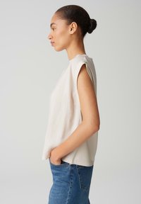 Helle beige ärmellose Bluse mit lockerer Passform, glatter Textur und dezenten Schulterdetails, kombiniert mit blauen Jeans.