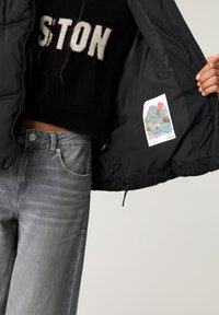 Sort pufferjakke med lynlås foran, der viser et vævet mærke og et farverigt landskabsgrafik. Sammen med grå jeans.