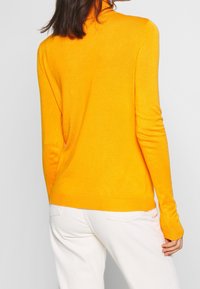 Jasnożółty sweter z długim rękawem i kołnierzem wysokim, wykonany z miękkiego materiału, z ściągaczami przy rękawach i dolnej krawędzi, noszony z białymi spodniami, pokazany od tyłu.