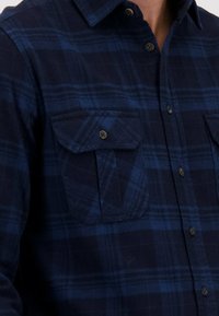 Donkerblauwe en zwarte geruite overhemden met klepborstzakken, gedragen door een persoon met gedeeltelijk zichtbare nek en kin.