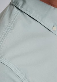 Camisa de botones azul claro con cuello doblado, textura de tejido suave y detalles de costura visibles a lo largo de las costuras.