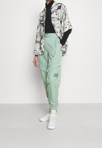 Mintgroene joggers met elastische manchetten en een logo-patch, gecombineerd met een licht shirt met patroon en een zwarte coltrui, gedragen met witte sneakers.