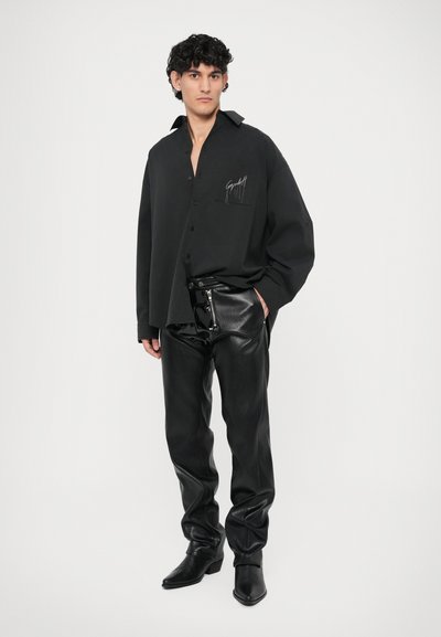 Schwarze, übergroße Button-Up-Bluse mit einer Tasche, versehen mit weißer Logo-Stickerei, kombiniert mit eng anliegenden schwarzen Lederhosen und spitzen Stiefeln.