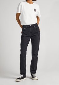 Jeans de mezclilla negra con corte recto, cinco bolsillos y cierre de botón, combinados con una camiseta blanca que presenta un pequeño logo.