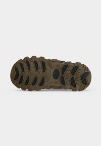 Semelle d’une chaussure de plein air robuste marron et noire avec des motifs de crampons profonds sur un fond gris uni.
