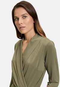 Blusa avvolgente verde oliva con scollatura a V profonda e maniche lunghe, realizzata in tessuto morbido con una leggera lucentezza. Presenta un dettaglio della tasca frontale.