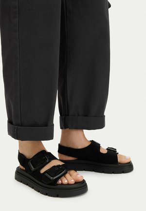 Feet wearing black chunky sandals with two large buckles, paired with black cuffed pants against a light background.